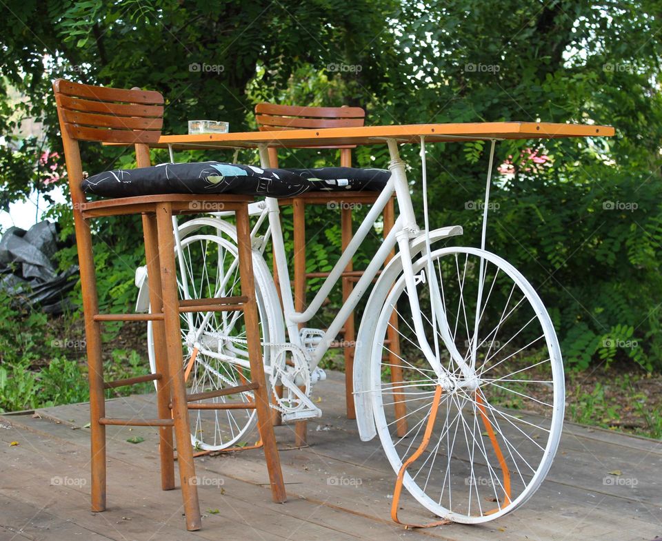 One lonely white bicycle with wooden board on top and two chairs.  Bicycle as a table