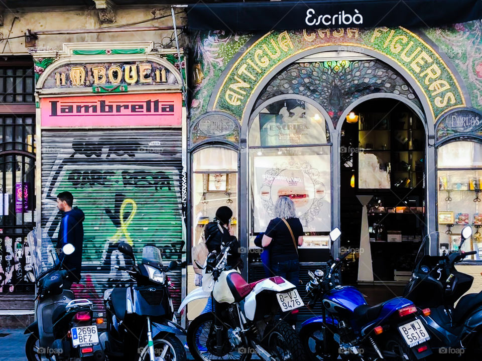 Street scene at La Rambla, Barcelona - Motorcycles are parked outside an antique store, next door displays a Lambretta sign and a spray painted yellow ribbon on the shutter