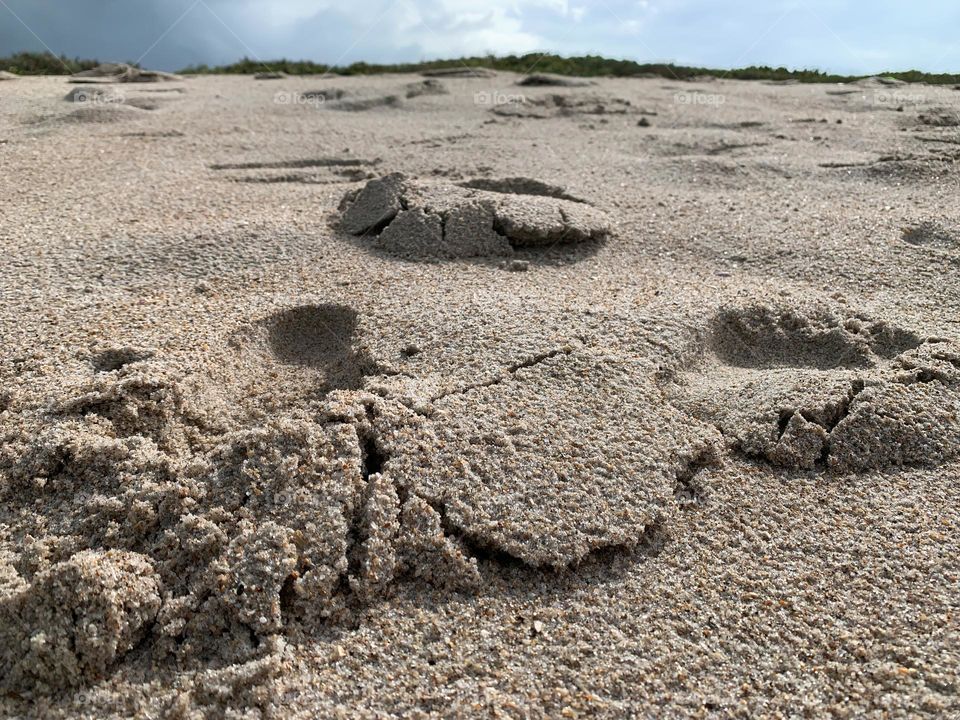 at the beach on the sand with multiple feet prints with hill of tropical plants and grey cloudy sky on the background.