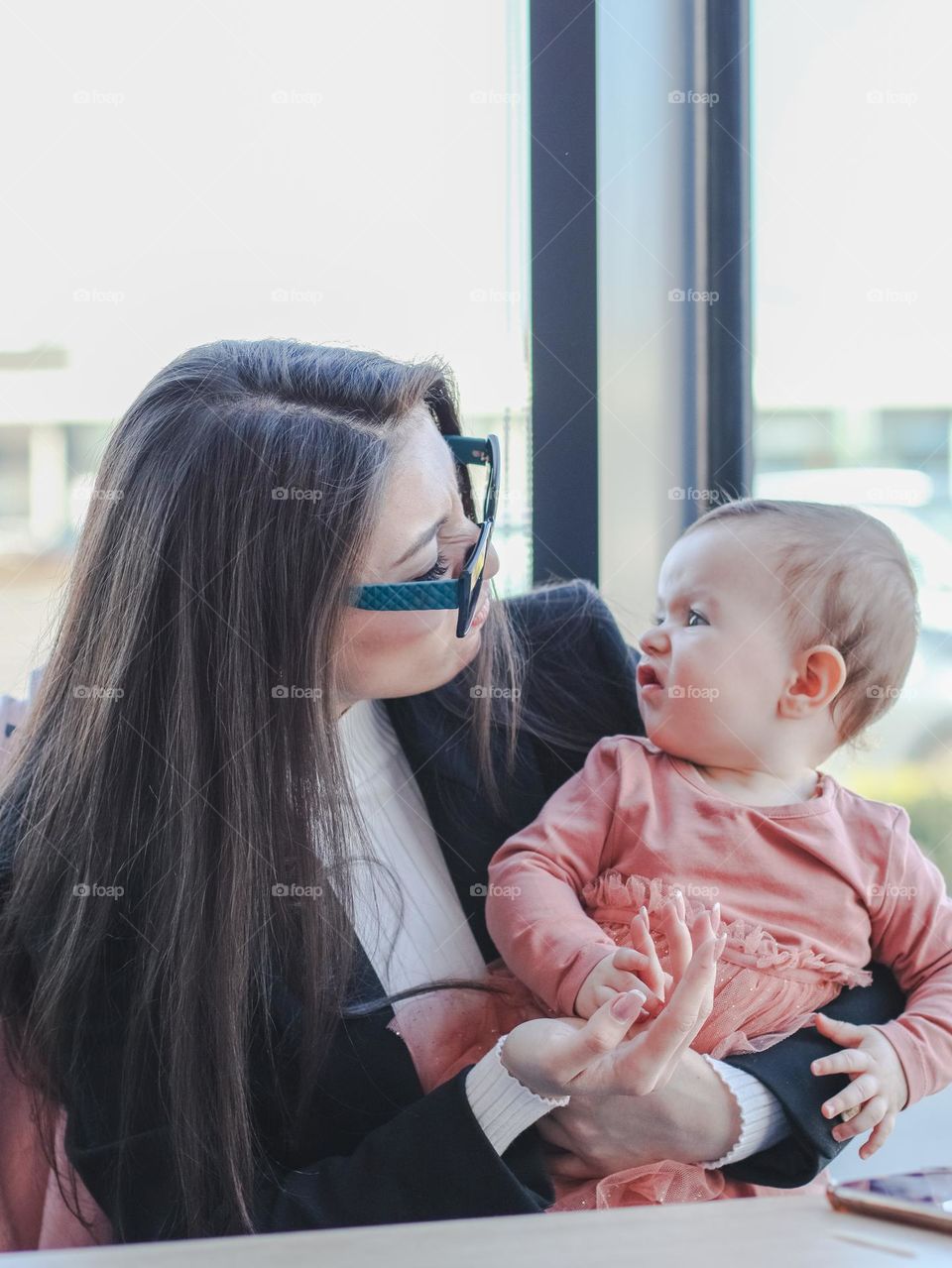 Portrait of a beautiful young business girl with a baby girl in her arms looking at her with an emotion of disgust while sitting at a glass table in a restaurant, close-up side view. People emotion concept, teaser game