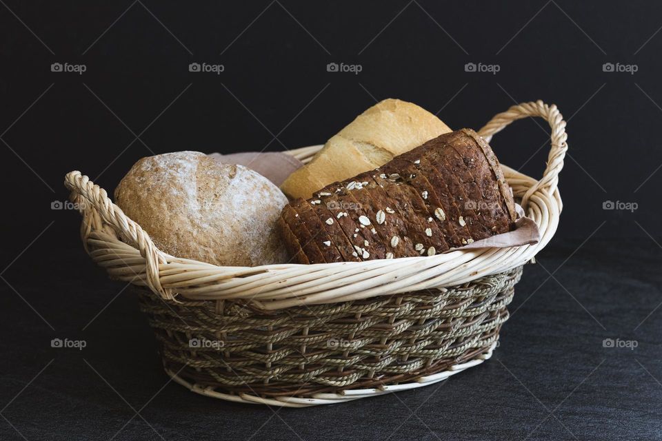 Three breads of different varieties in a wicker basket with handles lie in the center on a black stone table, close-up side view. The concept of baking bread.