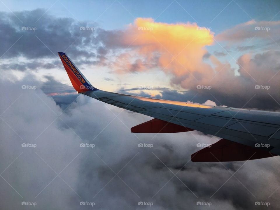 This photo captures the wing of a plane in flight amidst the clouds with a bit of sun peaking through.