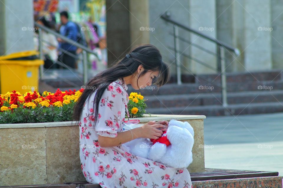 a girl in a white dress sits on a bench near a flower bed and holds a teddy bear in her hands