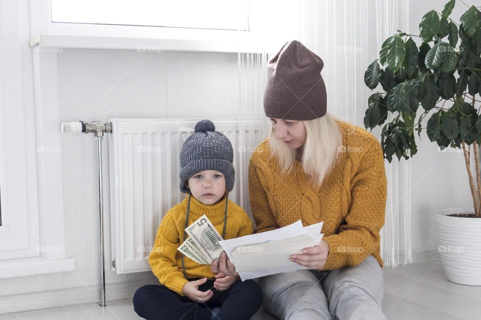 A mother with a small child in a yellow sweater and hats is counting money and thinking how to pay bills and taxes, near a heater with a thermostat.