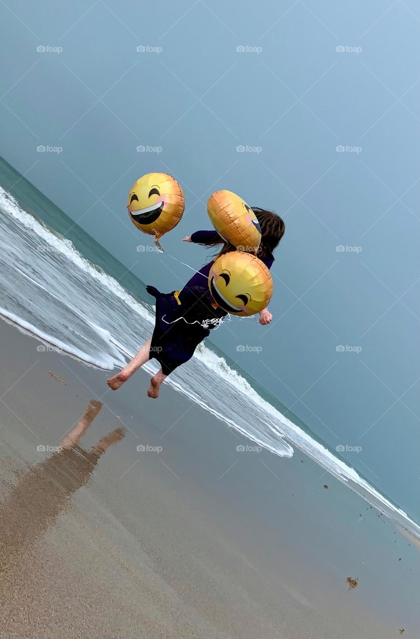 Little Girl Jumping And Running Having Fun With Helium Emoji’s Balloons On The Beach By The Ocean.