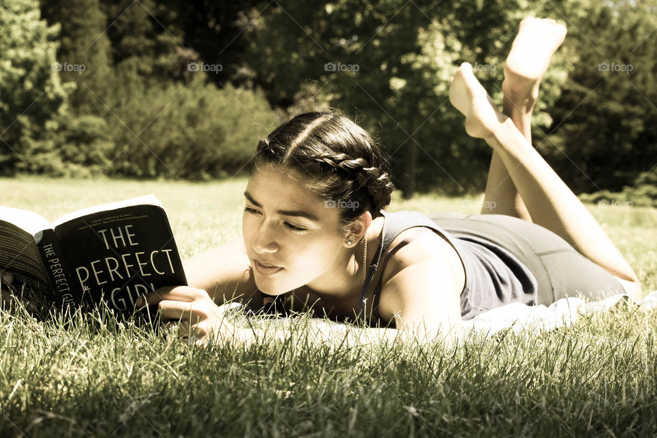 Teen girl is reading a book laying on the grass in a park