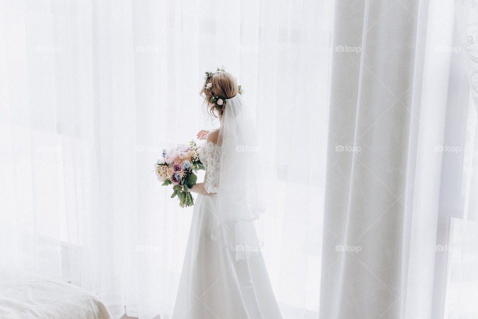 Bride in wedding dress with flowers bouquet near window