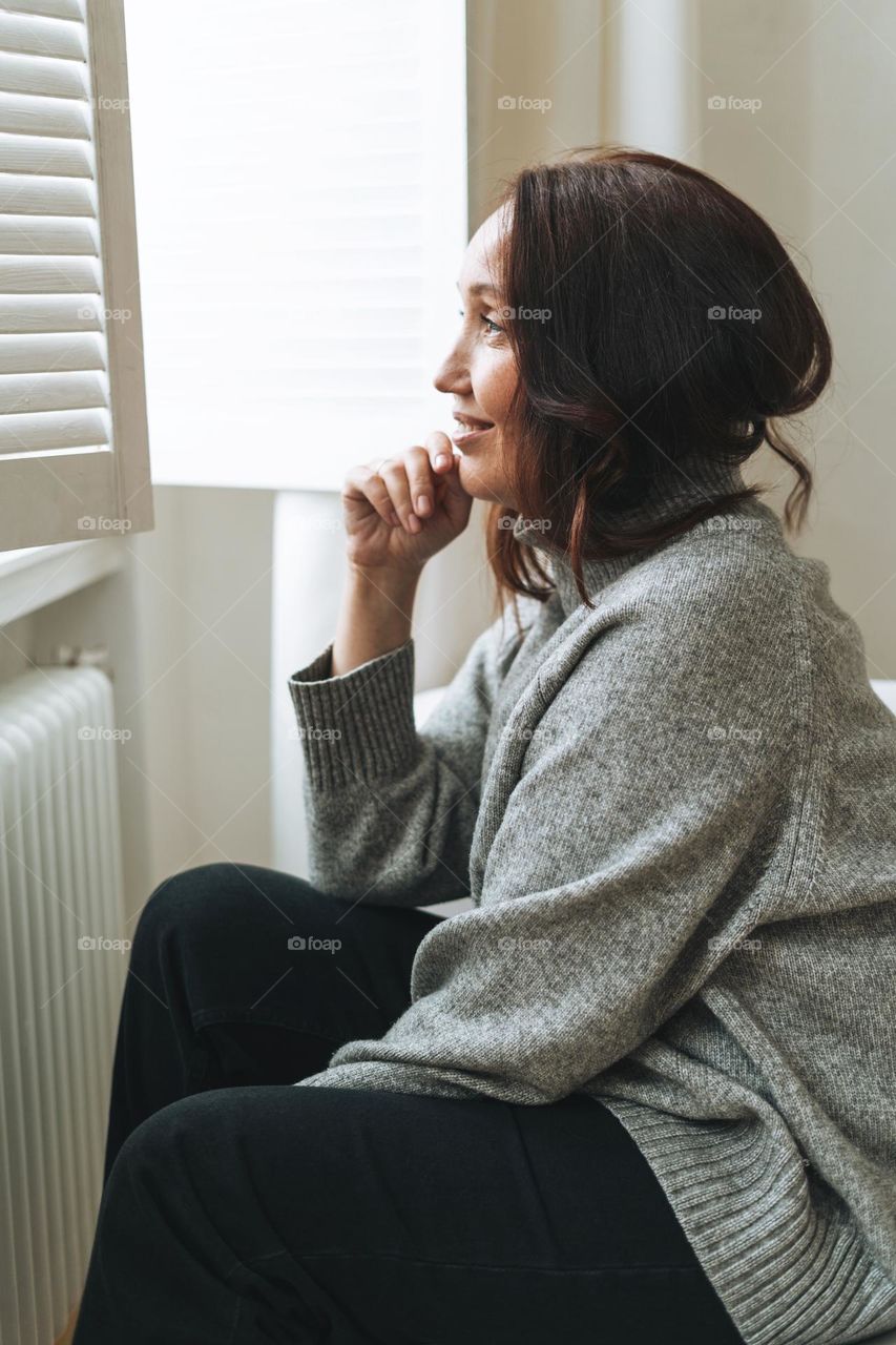 Young beautiful woman forty year with brunette hair in cozy knitted cardigan near window in bright interior at home