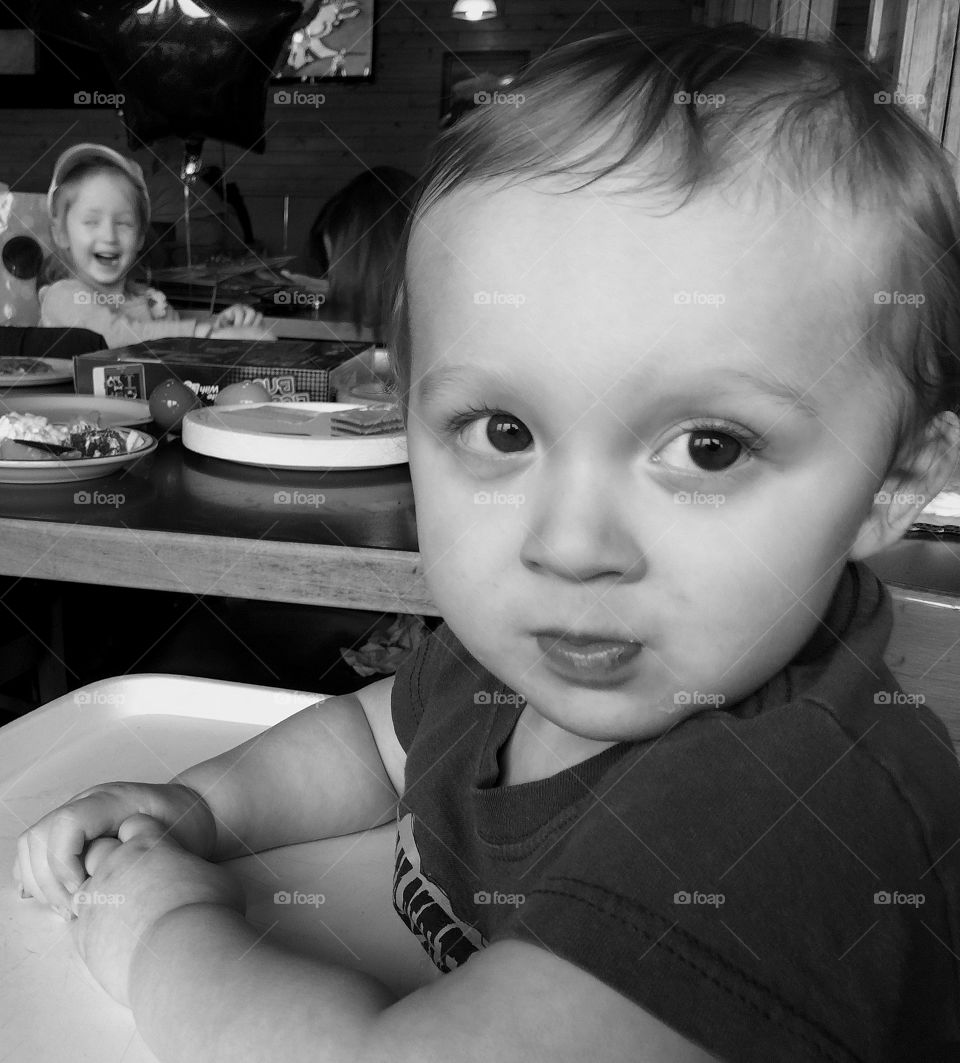 A toddler boy surprisingly waits patiently in a high chair for a piece of birthday cake. 