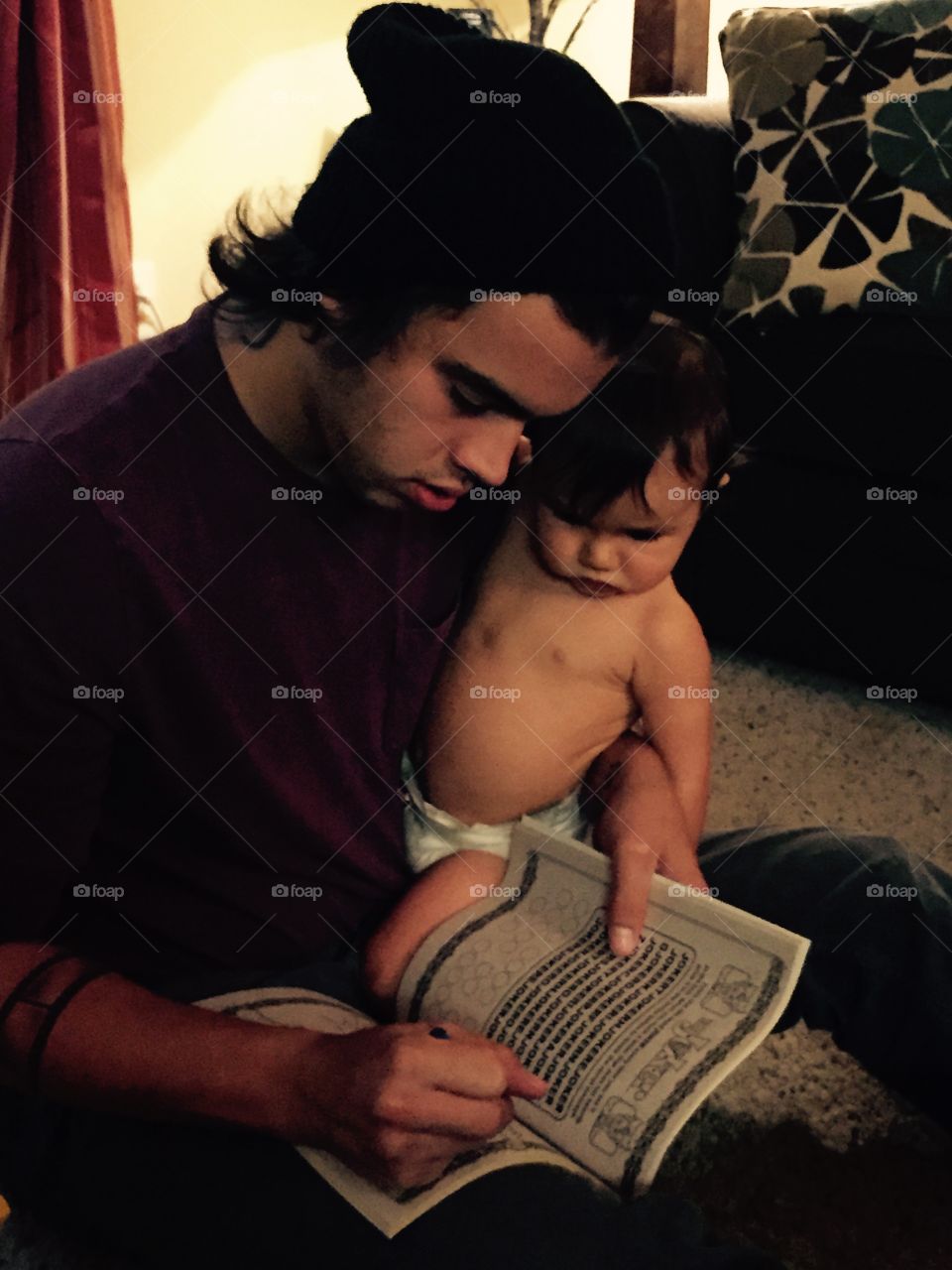 Young man and his baby boy inside home and dad is citing in color book. 