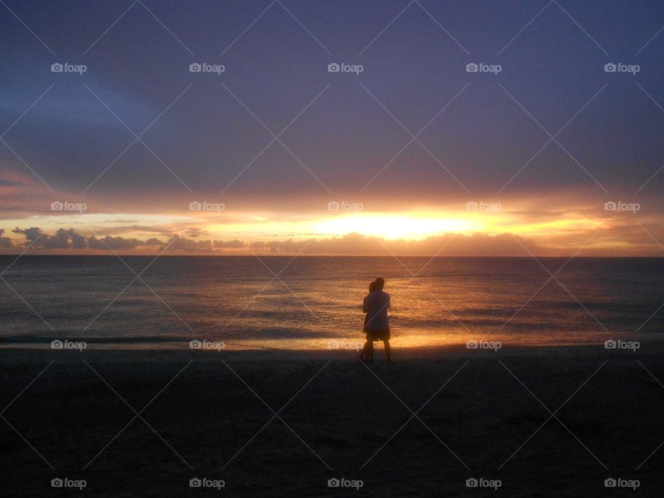 A person watches the sunset at the water’s edge on the beach. A streak of orange lights the sky as the sun disappears.