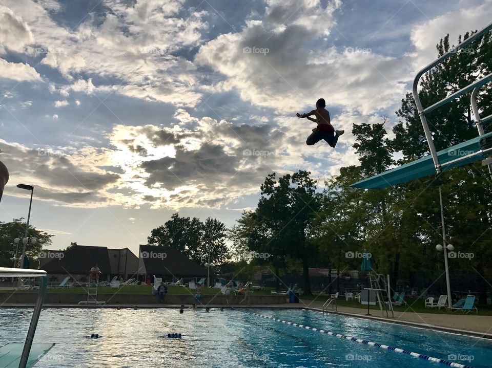 Boy jumping off high dive into swimming pool, sun hides behind clouds in the evening of a late summer day.