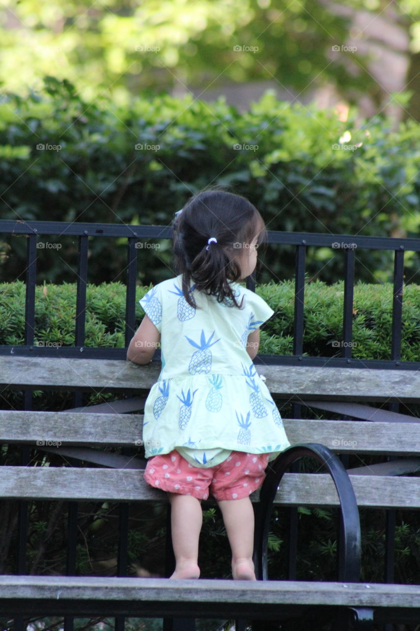 Toddler girl in green and blue pineapple shirt and pink pants standing tiptoe on wooden slatted bench 