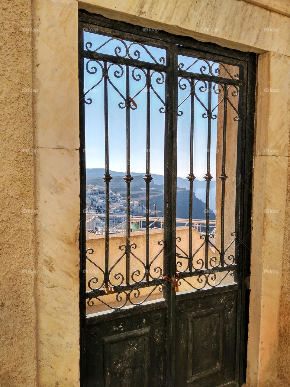 Simulation of a door with black iron bars in Santorini, Greece. Looking through the iron bars you can see in the background the Aegean Sea, some cliffs and a clear sky.