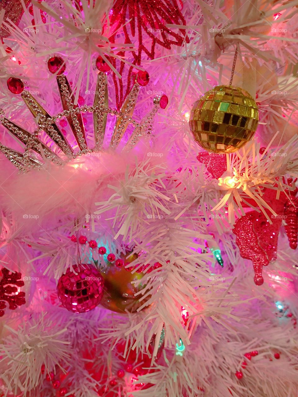 Beautiful gold and red ornaments hanging from a white Christmas tree with a pink glow. 