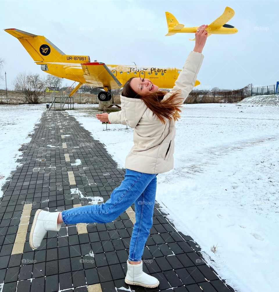 На фотографии изображена девушка позирующая перед ярким желтым самолетом . Она держит в руках миниатюрную копию того же самого самолета. Взгляд девушки наполнен восторгом, а её улыбка свидетельствует о радости и восхищении .