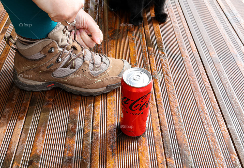 The metal friend follows but the furry one stays home! Coca cola zero can, New taste, Sweden. Woman’s hands tying laces to hiking boots with black cat paws in the background. Brown, wet wooden surface, sunny day