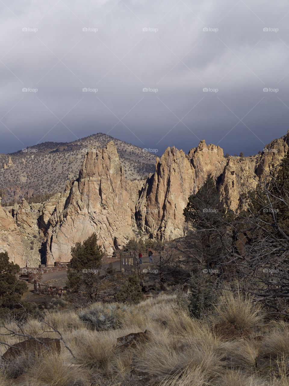 The jagged geology of Smith Rock State Park in Central Oregon with wild grasses and juniper trees in the foreground and stormy skies overhead on a winter day. 