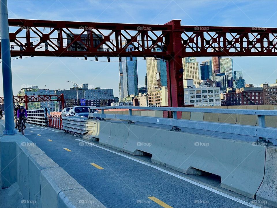 This is the “Pulaski Bridge” at “Newtown Creek” that connects “Greenpoint”, Bklyn to LIC in the direction that we are heading. Through the ornamental red metal gate we see nearby LIC high rises all aglow in the evening sun. 2024. Hypnotic Productions