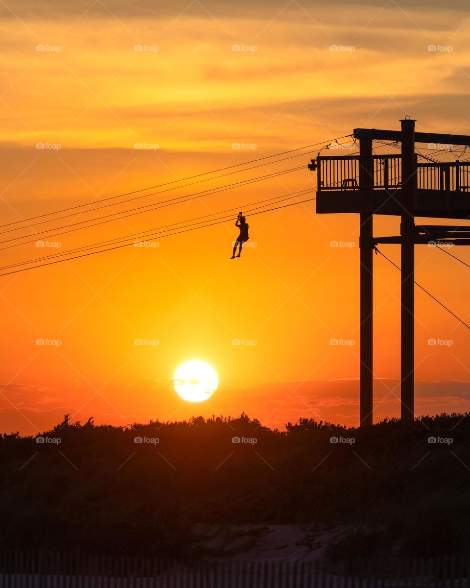 Silhouette of a person riding down on a zip line with a vibrant golden sunset in the background. 