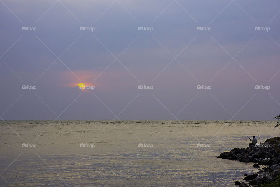 A man holding a fishing rod on the rock by the sea and a morning sunrise.