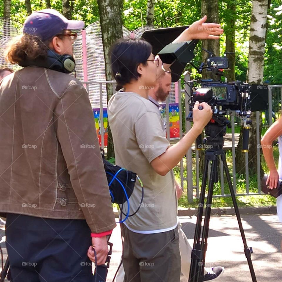 Summer day. Filming. Among the green trees, a film crew with professional equipment and a professional camera. A man who stands near a professional camera says something and gesticulates