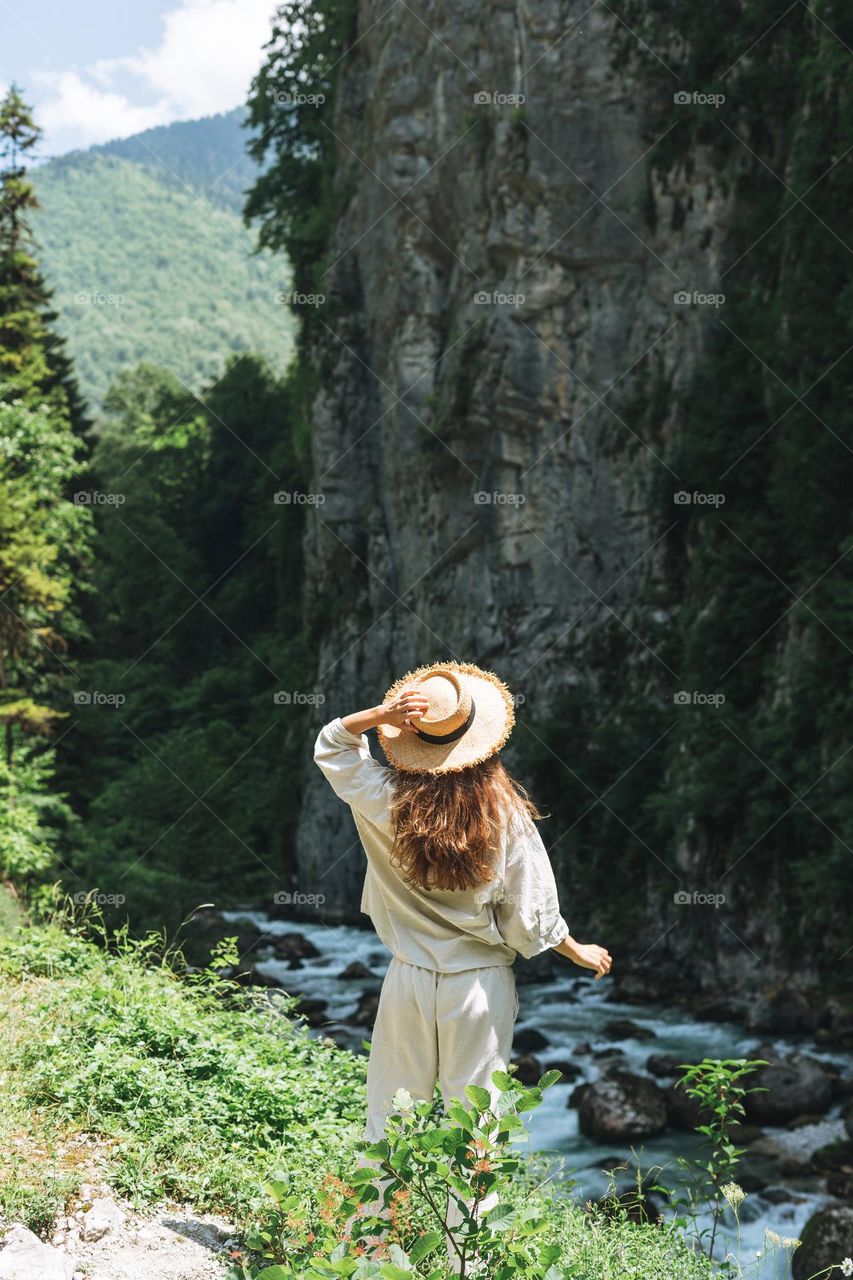 Young woman traveler with long blonde hair in straw hat looks at beautiful view of the mountain river, people from behind