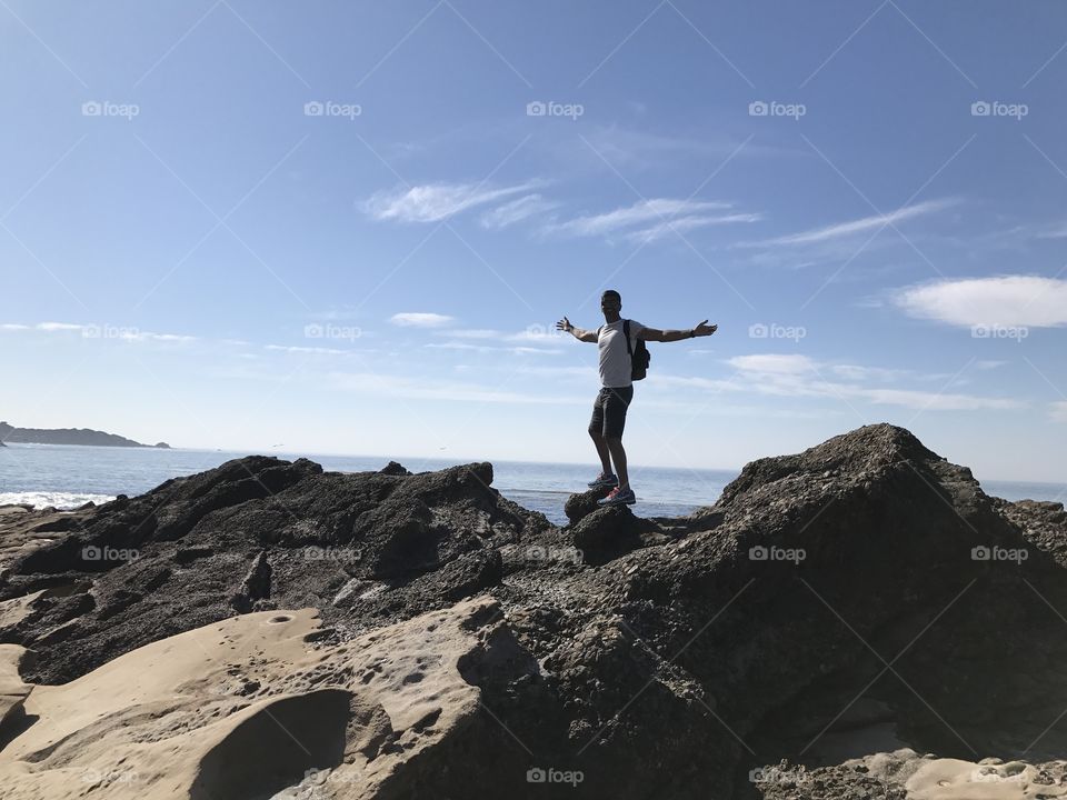 Man wearing shorts and a backpack adventuring standing high up on rock balancing with a bright blue sky in the background 