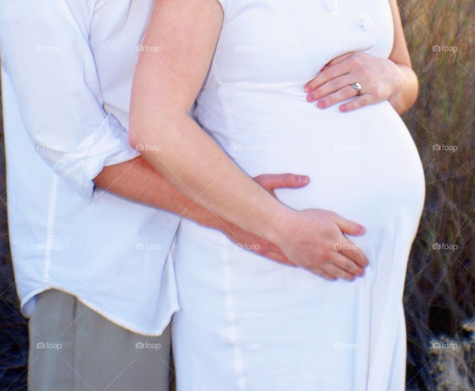 A husband and wife are eagerly awaiting the birth of their baby.  While the husband can't physically carry the child, his arms around his wife and the cradle of his wife's growing belly in his hands suggest a beautiful partnership.   
