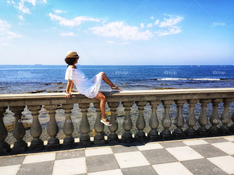 woman with hat dressed in white is sitting on a low wall on the Tyrrhenian sea in Livorno (Tuscany)