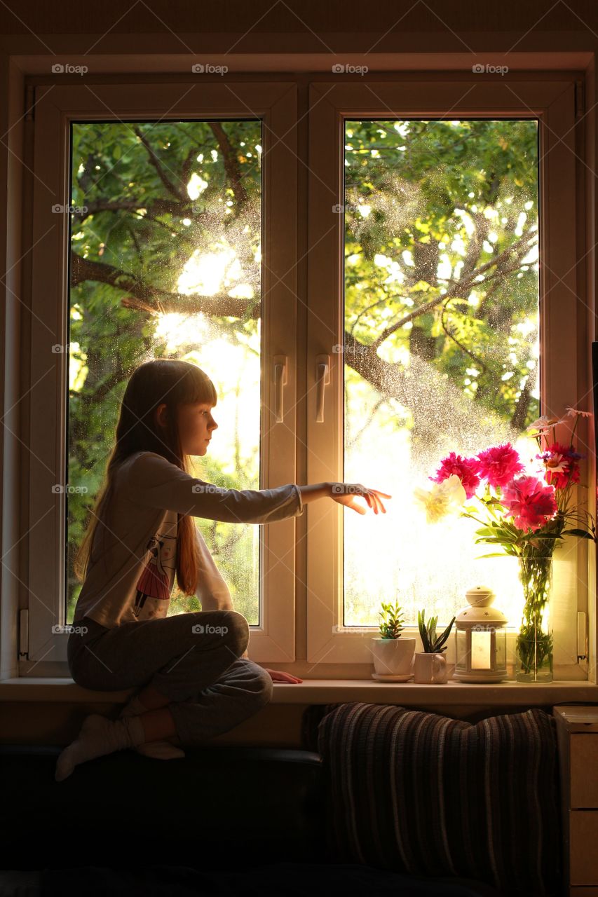 Little girl sitting on a windowsill reaches for flowers.