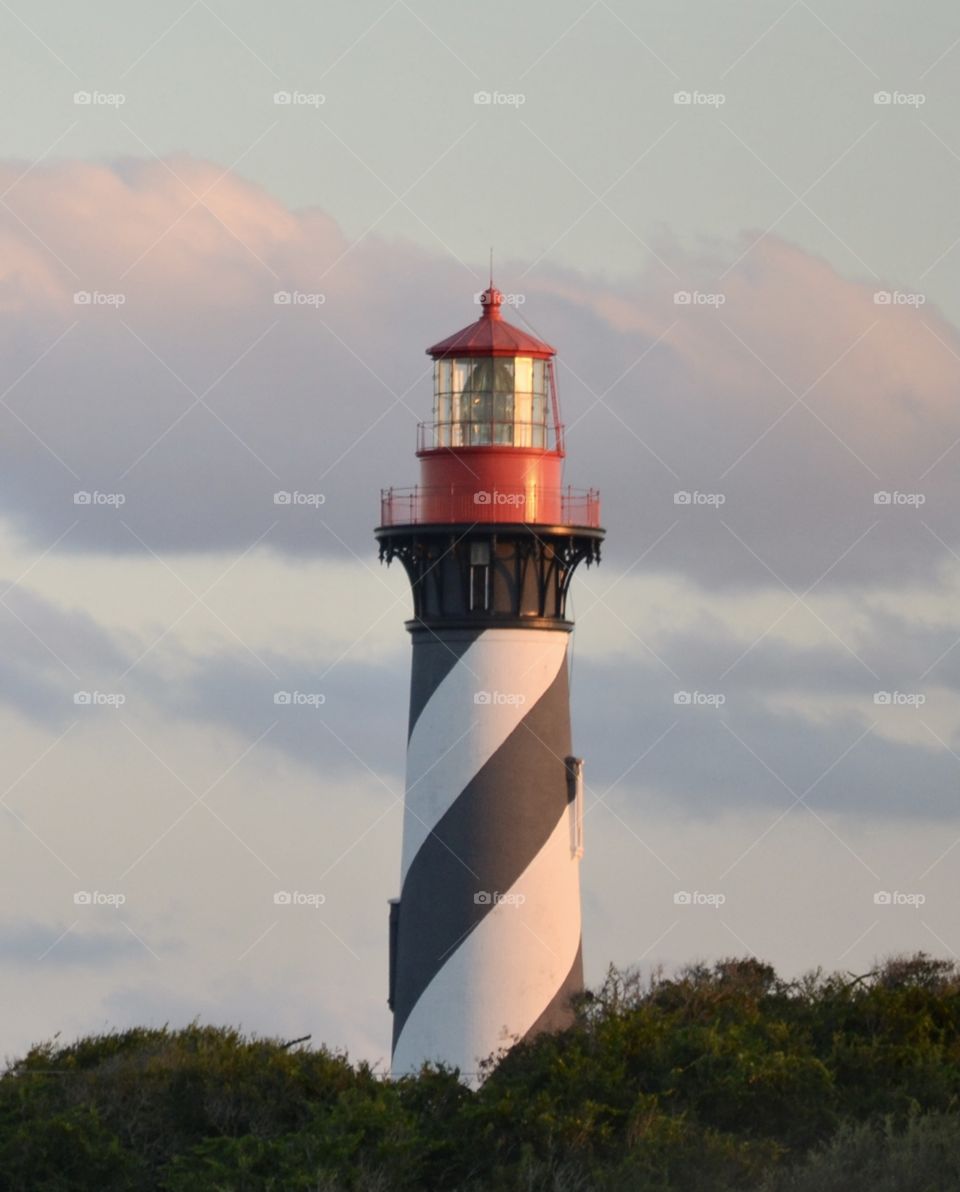 A black, white, and red lighthouse standing against a dusky blue, purple, and pink sky with green trees surrounding it