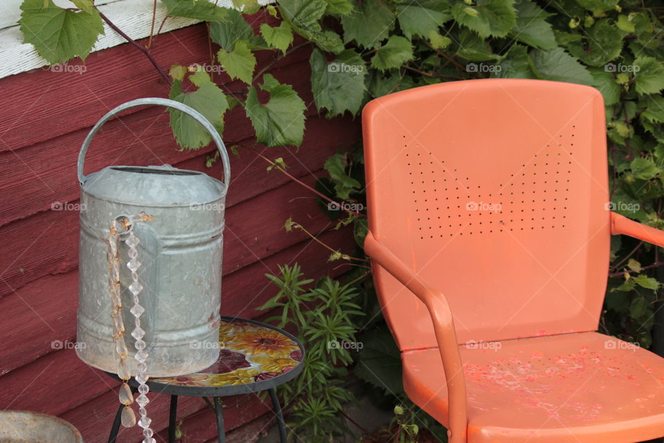 Decorative metal watering can with hanging plastic crystals on table beside orange rustic chair in yard