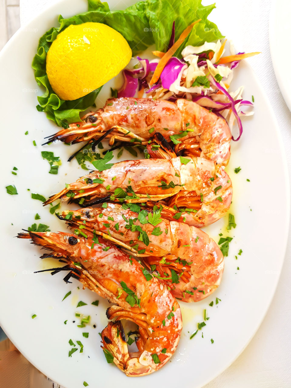 Top view to grilled shrimps sprinkled with chopped herbs next to a slice of lemon and green salad leaves on the plate.