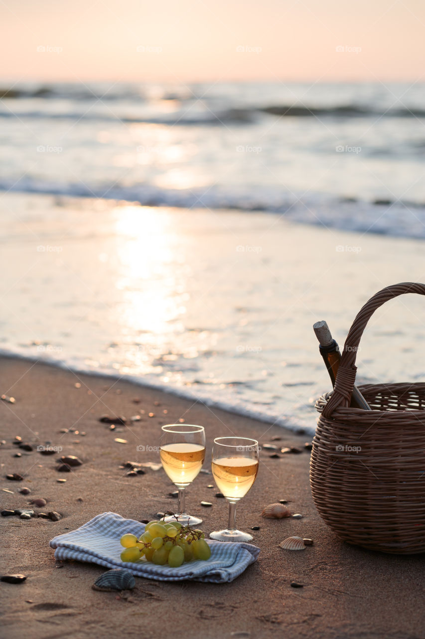 Two wine glasses with white wine standing on sand, on beach, beside grapes and wicker basket with bottle of wine