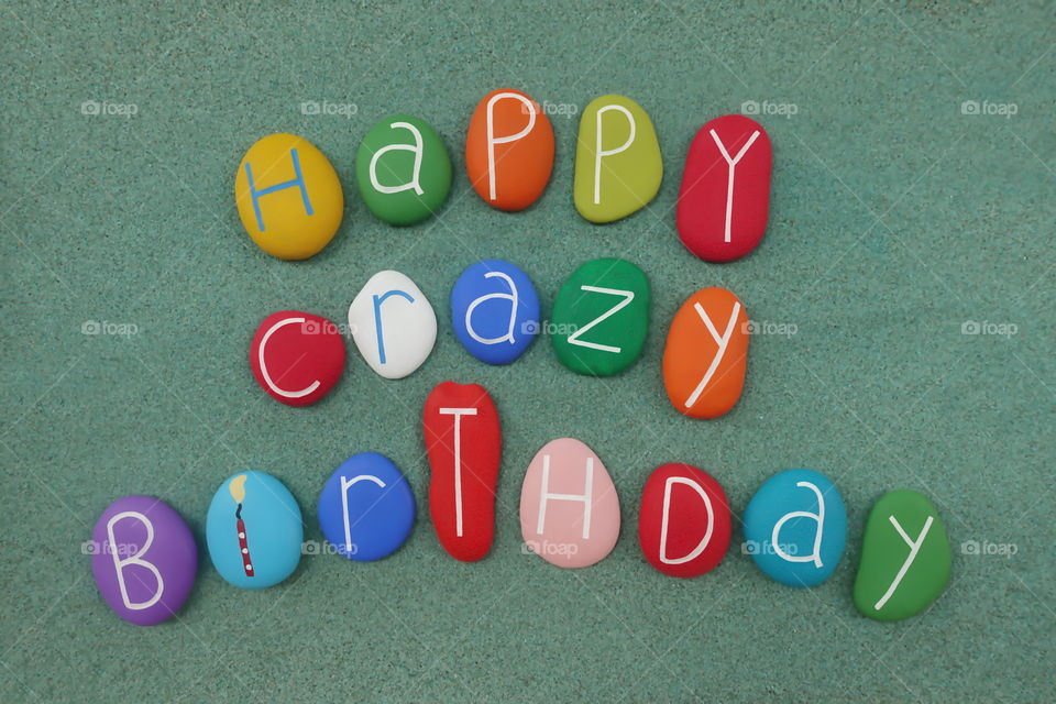 Happy Crazy Birthday text composed with colored stones over green sand 