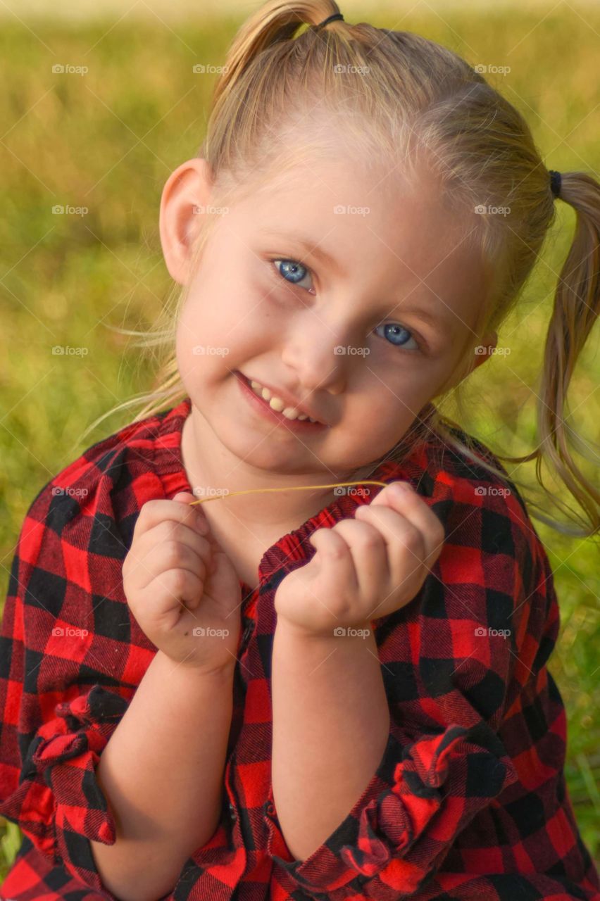 adorable blonde haired girl child with beautiful blue eyes and sweet smile wearing black and red checkered top with green grass in the background