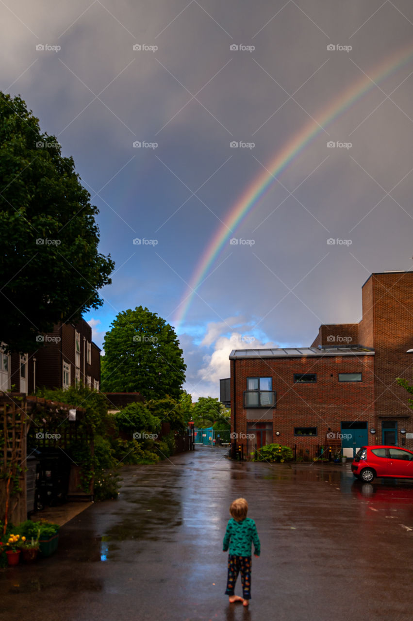 Small barefoot boy observes rainbow in the sky above buildings.