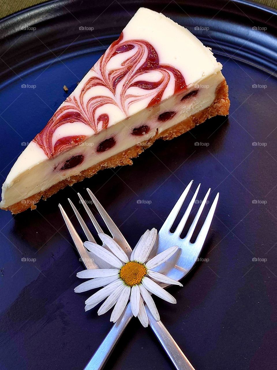 Food. On a black plate is a piece of cheese cake stuffed with blueberries. Near a piece of cake are two crossed forks, on the teeth of which lies a camomile