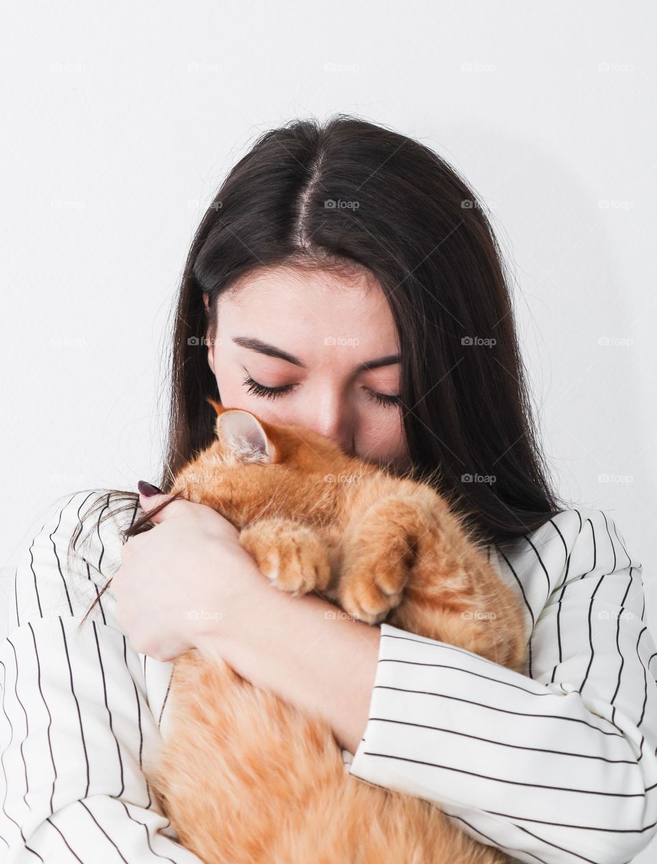 A beautiful young Caucasian brunette girl with long hair in a classic white jacket with black stripes holds a shy red thoroughbred cat in her arms, which hid its muzzle, side view close-up.