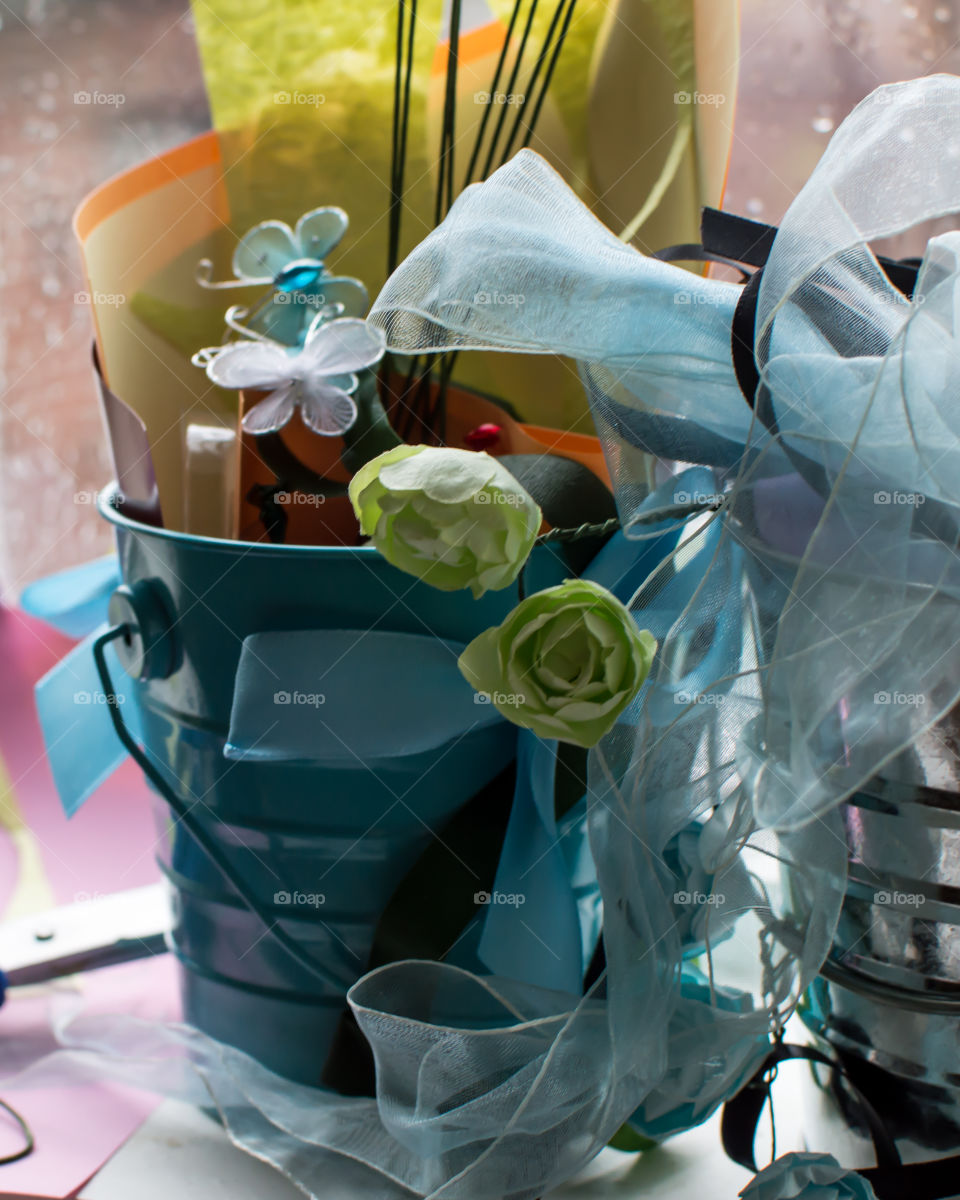 Different color and textured paper, flowers, butterfly and ribbon arts and craft materials in buckets on crafts table next to window on rainy day 