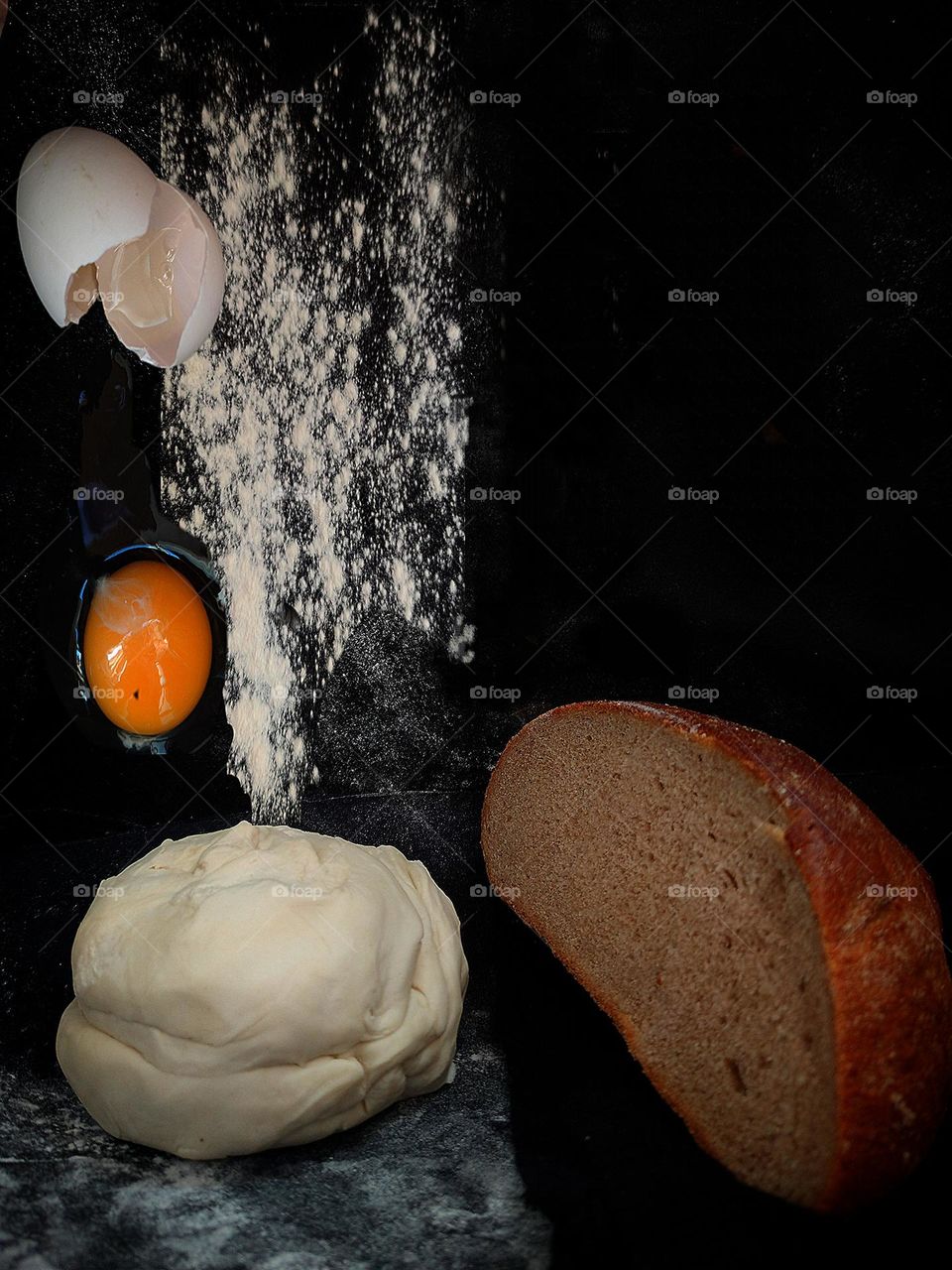 Homebaked bread. The black background is visually divided into two halves. On one half lies the dough, on which flour and a broken egg (dough ingredients) are sprinkled. Half of the baked bread lies on the second half background