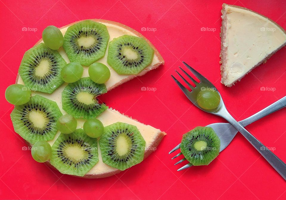Green color.Cheese cake topped with green kiwi slices and grapes.A triangular piece has been cut from the cake.Opposite the empty space are two crossed metal forks with a grape and a kiwi slice.In the upper corner lies a triangle white of cheese cake