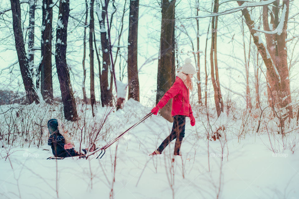 Teenage girl pulling sled with her little sister, a few years old girl, through forest covered by snow while snow falling, enjoying wintertime, spending time together. Girls are wearing winter clothes