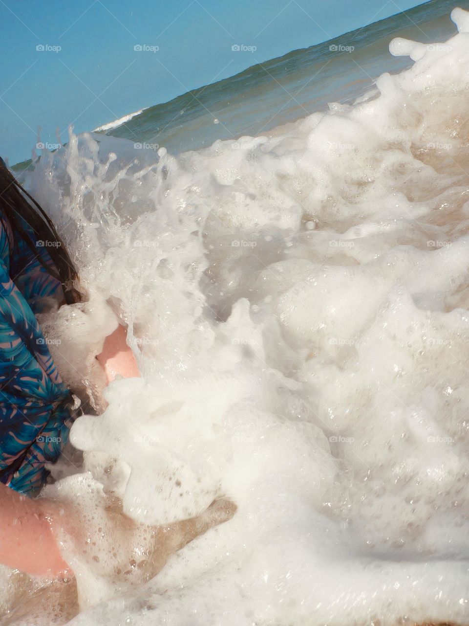 Beach vacation splash from the salty waves on the child girl dressed in a blue modest swimsuit at the Atlantic Ocean sitting on the seashore on the sand with hands playing in the water.