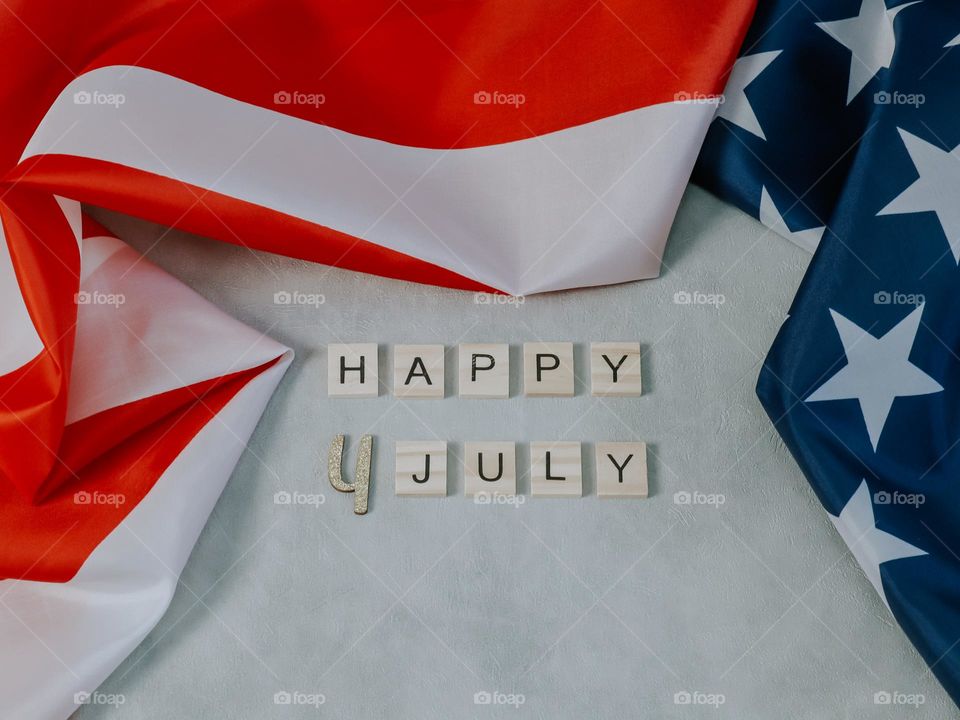 An American flag with wooden cubes with the words Happy 4th of July lying on a light-colored concrete background, close-up side view.