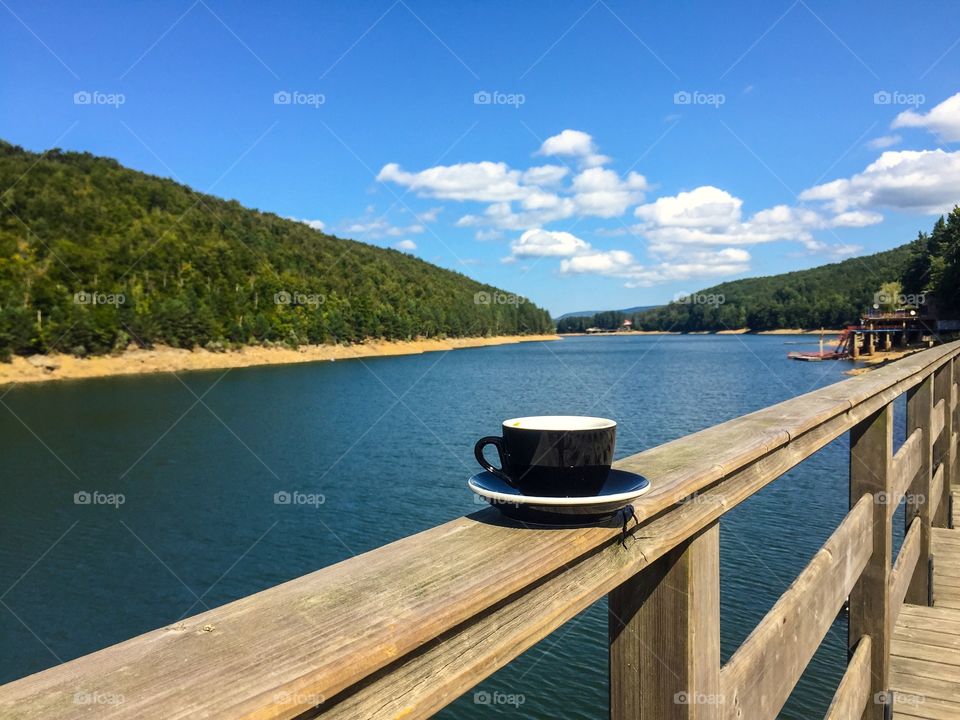 Cup of coffe on plate served on a terrace near a lake surrounded by forest