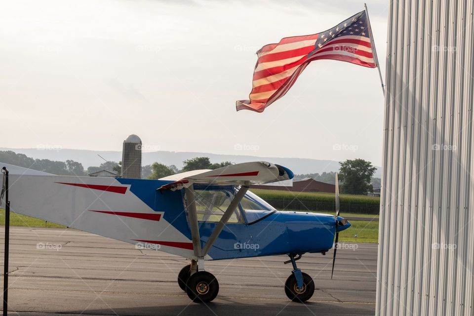 Foap, American Flag: A brilliant American Flag flaps in the breeze over a red, white, and blue two-seater airplane on the tarmac. 