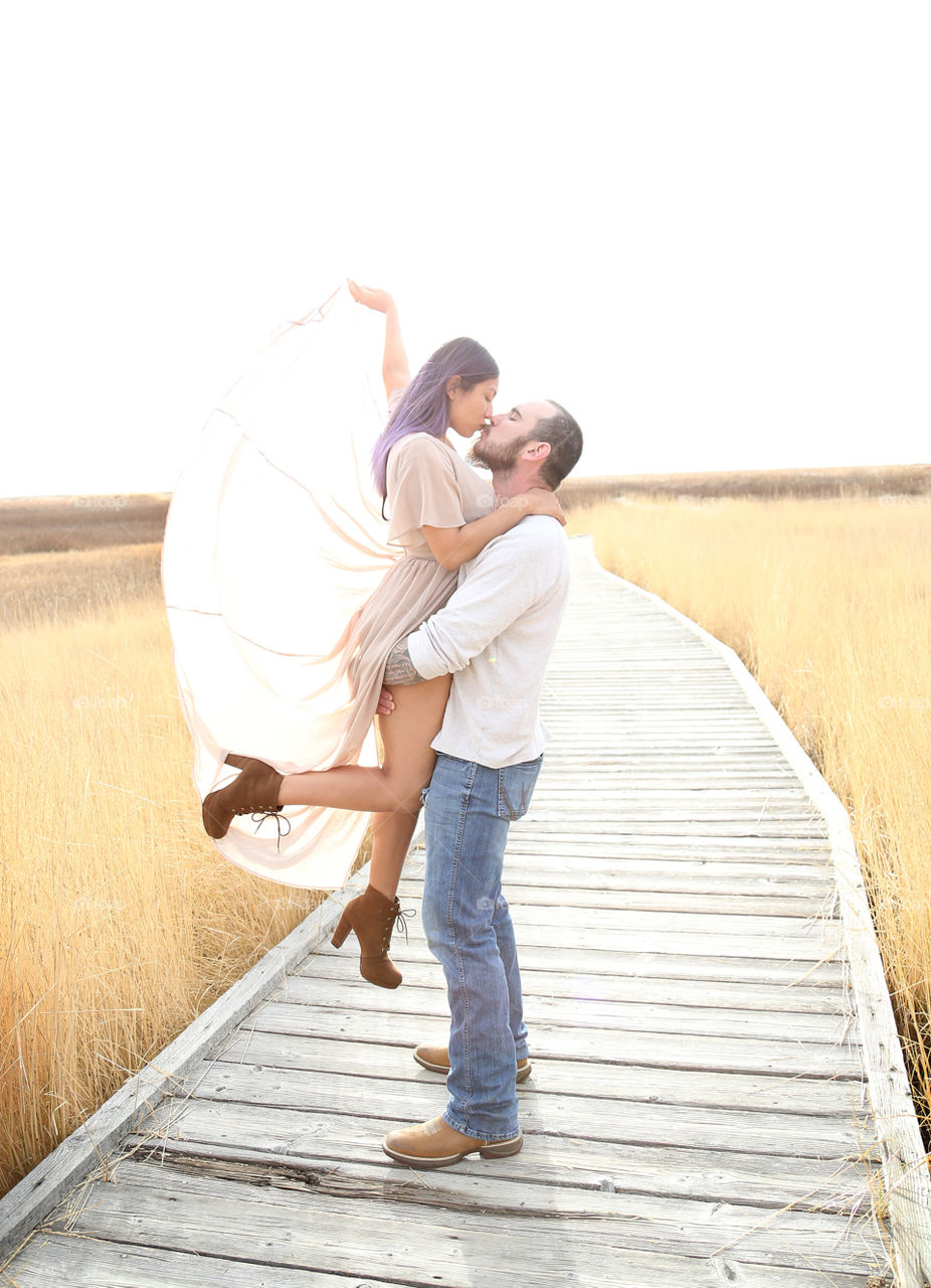 Man and woman couple in love standing on wooden plank walkway in golden wheat straw field. Man is wearing blue denim jeans, cowboy boots, off white sweater. Woman wearing boots, mauve dress with wings. Woman is held in air with arm outstretched.