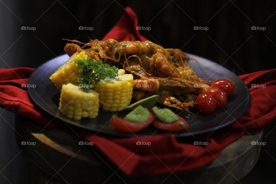 Asian food balado fried prawns with corn chunks, lemon slices and tomatoes served on black plate with red cloth bottom and wooden coaster. Asian style street seafood.